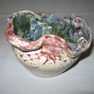 May include: A handmade ceramic bowl with a unique, wavy rim and a speckled glaze in shades of white, brown, red, blue, and green.
