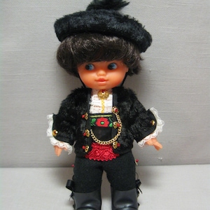 May include: A vintage doll dressed in traditional attire. The doll wears a black hat, jacket with gold trim, black pants, and boots. The jacket has red and green floral embroidery and a gold chain. The doll's outfit includes a red belt and white lace cuffs.