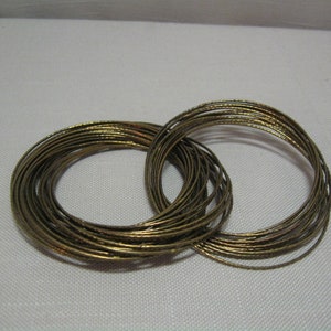 May include: A set of gold-toned metal bangles, stacked and intertwined. The bangles are thin and have a slightly textured surface.