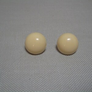 Op de afbeelding: Twee ivoorkleurige, bolvormige objecten, mogelijk kralen of decoratieve elementen, zijn geplaatst op een gestructureerd, lichtgrijs oppervlak. De bollen hebben een glanzende afwerking en zijn gelijkmatig verdeeld.