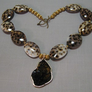 May include: A necklace made of oval-shaped brown and white shell beads with gold spacers. The necklace has a large, irregular-shaped pendant made of black stone with a gold rim.