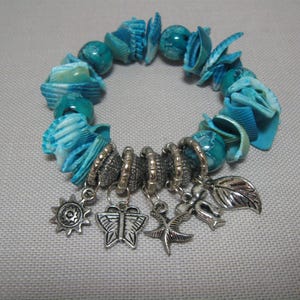 Puede incluir: Un brazalete turquesa y plateado con una variedad de encantos, incluyendo una mariposa, un sol, un pájaro y una hoja. El brazalete está hecho de piezas de concha azul y blanca.