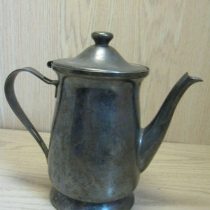 Puede incluir: Una tetera de plata con cuerpo redondeado y tapa. La tetera tiene un asa y un pico.