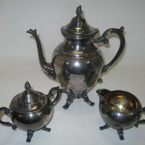 Op de afbeelding: Een zilveren theeservies met een theepot, roomkan en suikerpot. De set heeft een traditioneel ontwerp met sierlijke details en drie poten.