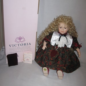 May include: A porcelain doll with blonde curly hair wearing a red and black floral dress with a white lace collar and a red heart. The doll is sitting on a white surface with a box and a small velvet bag in the background. The box has the text "Step into the world of Victoria" and "Victoria's Collectibles" printed on it.
