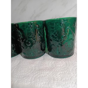 May include: A set of three emerald green glass tumblers featuring a raised, embossed holiday-themed pattern. The design includes visible motifs of candy canes, stockings, and festive ornaments moulded directly into the exterior surface of the glass.