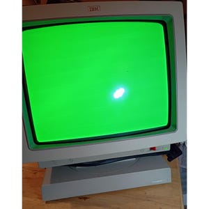 May include: A vintage IBM computer monitor featuring a light gray plastic casing and a bright, solid green cathode ray tube display screen. The monitor includes a prominent IBM logo at the top center and a small red power switch on the front panel.