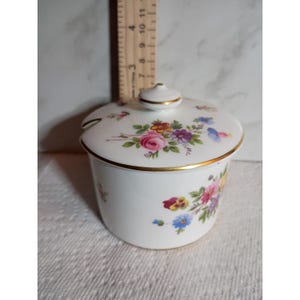 May include: A white porcelain sugar bowl with a lid featuring a colourful floral pattern of pink roses, purple, yellow, and blue flowers. The rim and lid knob are trimmed in gold. A wooden ruler shows the height is approximately 9 cm.