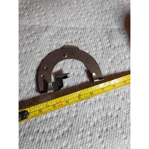 May include: A semi-circular silver-toned metal component featuring two small protruding hooks at the base and two circular holes along the curved edge. A yellow measuring tape is positioned alongside, indicating a width of approximately 3.5 inches.