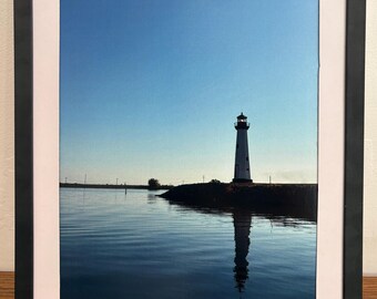 May include: Framed print showing a white lighthouse with a black top, reflected in still water. The sky is clear blue. The frame is black with a white mount. The scene evokes a sense of peace.