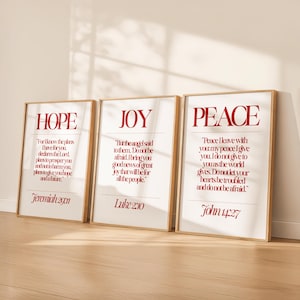May include: Three framed art prints with the words "HOPE", "JOY", and "PEACE" in red lettering. Each print features a quote from the Bible. The frames are a light wood color, and the background is white.