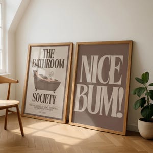 May include: Two framed art prints. One print has the text "THE BATHROOM SOCIETY" with an illustration of a person in a bathtub. The other print says "NICE BUM!" in large white letters on a brown background.