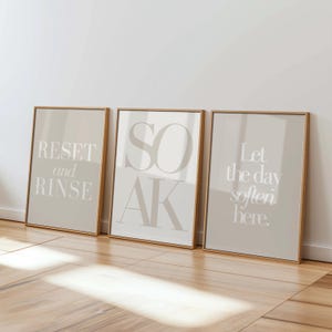 以下が含まれることがあります： 「RESET and RINSE」、「SOAK」、および「Let the day soften here」というテキストが書かれた3つの額入りアートプリント。プリントは、ベージュの背景と明るいテキストを備えたニュートラルなカラーパレットで、木製のフレームに収められています。