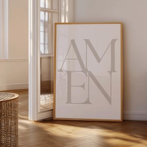Może przedstawiać: Oprawiony wydruk z napisem "AMEN" w dużych, stylizowanych, szarych literach. Grafika umieszczona jest na jasnym tle i oprawiona w naturalnym odcieniu drewna. Wydruk eksponowany jest w jasnym pomieszczeniu z drewnianą podłogą.