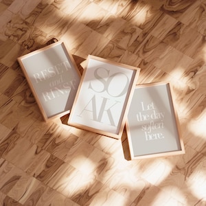 May include: Three framed prints with light gray backgrounds and wooden frames are displayed on a wooden floor. The prints feature the words "RESET and RINSE", "SOAK", and "Let the day soften here" in a light gray font.