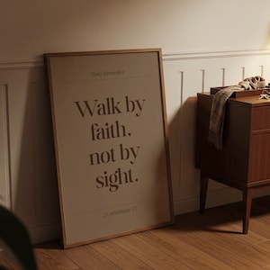May include: A framed poster with the text "Walk by faith, not by sight" in brown lettering. The poster is leaning against a white wall and wooden floor. A wooden cabinet is to the right of the poster.
