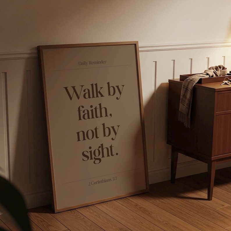 May include: A framed poster with the text "Walk by faith, not by sight" in brown lettering. The poster is leaning against a white wall and wooden floor. A wooden cabinet is to the right of the poster.