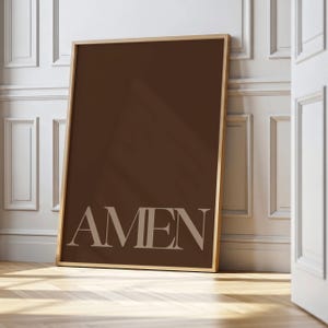 May include: A framed art print featuring the word "AMEN" in a light beige font against a dark brown background. The frame is a light wood color. The artwork is leaning against a white wall.