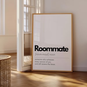 May include: A framed print with the word "Roommate" in bold black letters. Below, the definition reads: "someone who witnesses every version of you and still renews the lease." The frame is a light brown color.