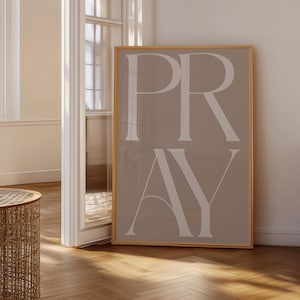 Może przedstawiać: Oprawiony wydruk artystyczny ze słowem "PRAY" w dużych, eleganckich, białych literach na tle w kolorze taupe. Rama jest w ciepłym, jasnobrązowym kolorze. Grafika jest eksponowana w pomieszczeniu z drewnianą podłogą.