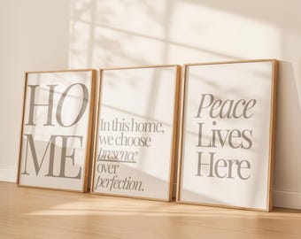 May include: Three framed art prints with light gray text on a white background. The prints read "HOME", "In this home, we choose presence over perfection.", and "Peace Lives Here". The frames are a light wood color.