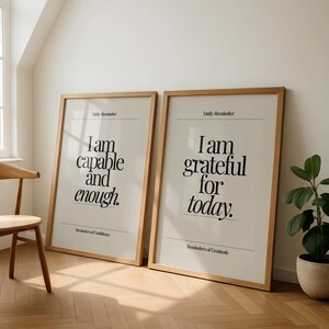 May include: Two framed posters with inspirational quotes. The left poster reads "I am capable and enough." The right poster says "I am grateful for today." Both have a light beige background, black text, and wooden frames. The posters are leaning against a white wall.