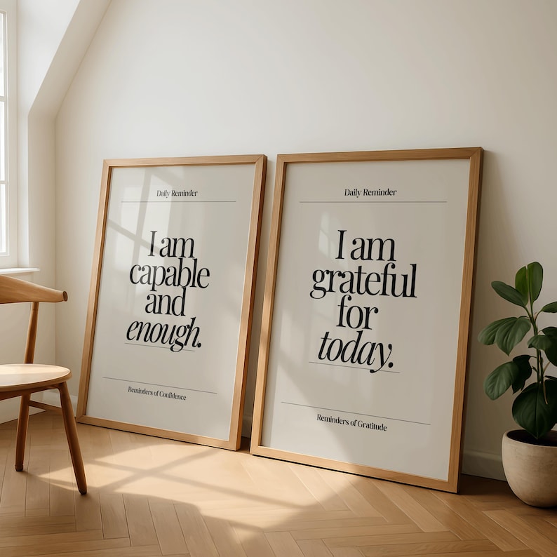 May include: Two framed posters with inspirational quotes. The left poster reads "I am capable and enough." The right poster says "I am grateful for today." Both have a light beige background, black text, and wooden frames. The posters are leaning against a white wall.