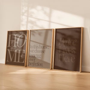 May include: Three framed art prints with gold frames. The prints feature text: "HOME", "In this home, we choose presence over perfection.", and "Peace Lives Here". The prints are in shades of brown and beige.