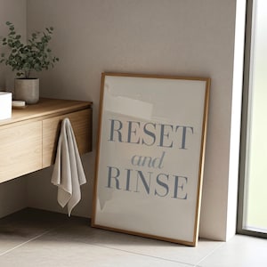 May include: A framed print with the words "RESET and RINSE" in blue lettering. The print is leaning against a wall in a bathroom setting, with a wooden vanity and a plant in the background. A gray towel hangs from the vanity.