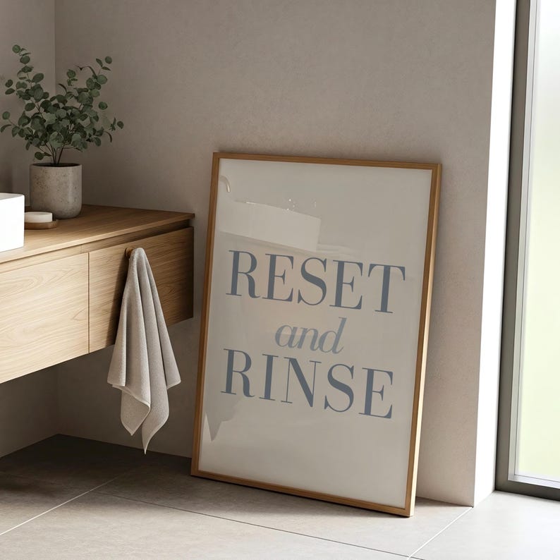 May include: A framed print with the words "RESET and RINSE" in blue lettering. The print is leaning against a wall in a bathroom setting, with a wooden vanity and a plant in the background. A gray towel hangs from the vanity.