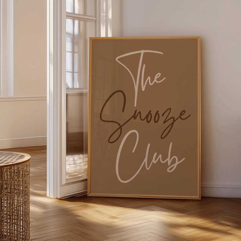Pode incluir: Uma impress&atilde;o de arte emoldurada com as palavras "The Snooze Club" em uma fonte de script elegante. O texto &eacute; branco e marrom sobre um fundo bege. A moldura &eacute; de cor marrom claro. A impress&atilde;o &eacute; exibida em uma sala clara.