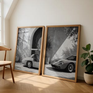 May include: Two framed black and white wall art prints featuring classic sports cars. The left print shows a vintage car with gull-wing doors open, while the right print displays a low-profile racing car with dual stripes parked near a stone wall.