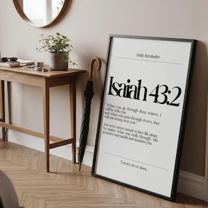 K&ouml;nnte beinhalten: Ein gerahmter Druck mit dem Text "Isaiah 43:2" in Schwarz auf wei&szlig;em Hintergrund. Der Druck befindet sich in einem schwarzen Rahmen und lehnt an einer Wand. Ein Holztisch, eine Topfpflanze und ein Regenschirm sind ebenfalls zu sehen.