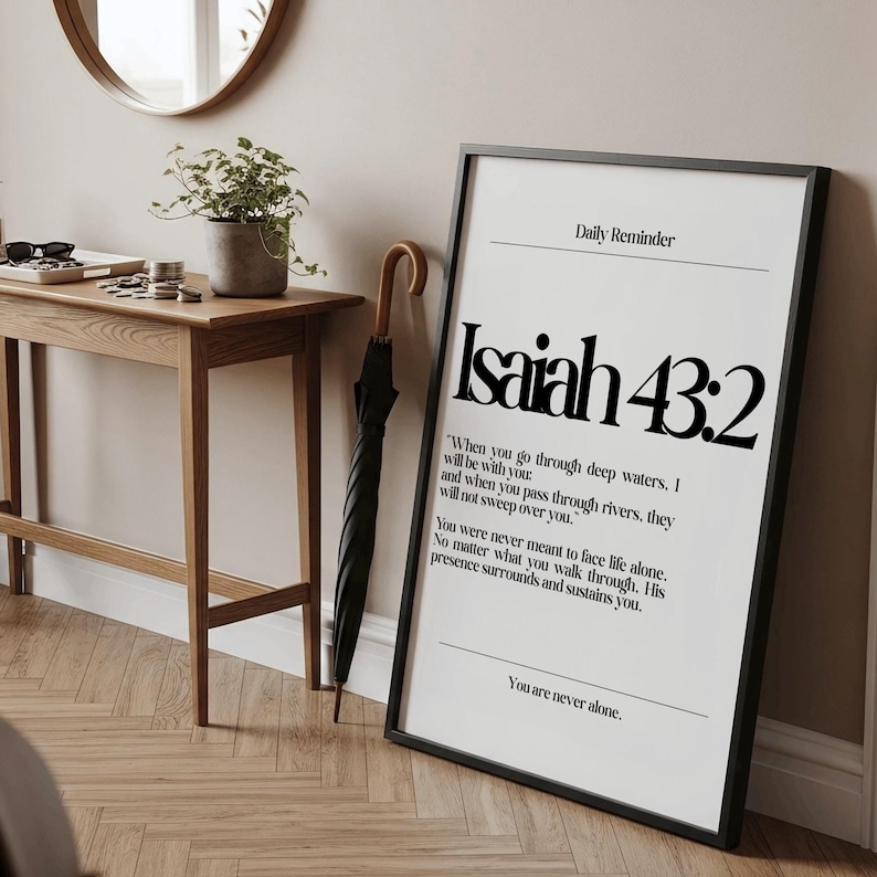 K&ouml;nnte beinhalten: Ein gerahmter Druck mit dem Text "Isaiah 43:2" in Schwarz auf wei&szlig;em Hintergrund. Der Druck befindet sich in einem schwarzen Rahmen und lehnt an einer Wand. Ein Holztisch, eine Topfpflanze und ein Regenschirm sind ebenfalls zu sehen.