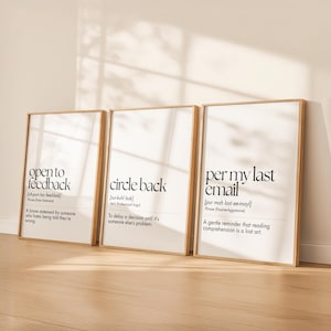 May include: Three framed art prints with gold-colored frames lean against a wall. Each print features black text on a white background. The phrases are "open to feedback", "circle back", and "per my last email".