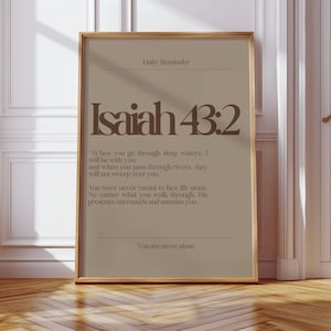 May include: A framed print with the text "Isaiah 43:2" in a stylish font. The print has a beige background with the words "Daily Reminder" at the top and a quote below. The frame is a warm gold color.