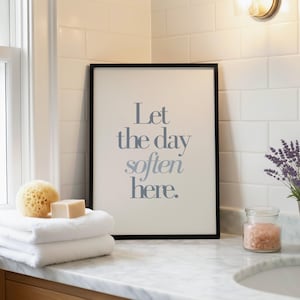 May include: A framed print with the text "Let the day soften here" in blue-gray lettering. The print is displayed on a marble countertop with white towels, a natural sponge, soap, and a jar of pink bath salts. A sprig of lavender is in a small pot.