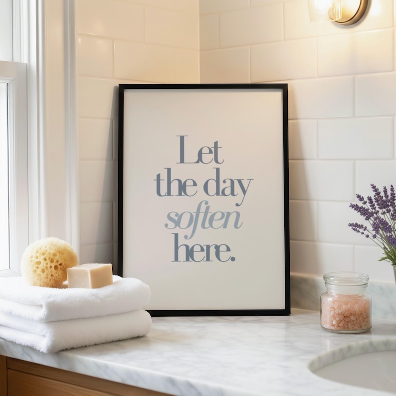 May include: A framed print with the text "Let the day soften here" in blue-gray lettering. The print is displayed on a marble countertop with white towels, a natural sponge, soap, and a jar of pink bath salts. A sprig of lavender is in a small pot.
