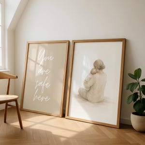 Könnte beinhalten: Zwei gerahmte Kunstdrucke. Einer zeigt den Text "You are safe here" in weißer Schrift auf beigem Hintergrund. Der andere zeigt eine Mutter, die ein Baby in einer weichen, neutralen Farbpalette hält. Beide sind in hellen Holzrahmen.