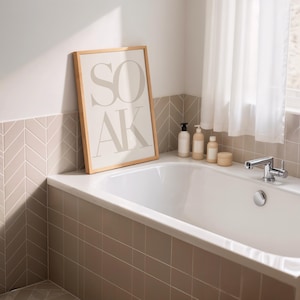 May include: A bathroom scene featuring a white bathtub, a framed print with the word "SOAK", and various bath products. The print has a light wood frame and the text is in a soft gray color. The walls are tiled in a neutral color.