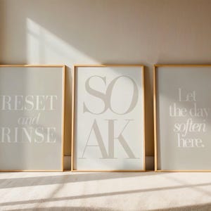 May include: Three framed wall art prints with inspirational text. The prints are in neutral tones of beige and white, with gold frames. The text on the prints reads "RESET and RINSE", "SOAK", and "Let the day soften here."