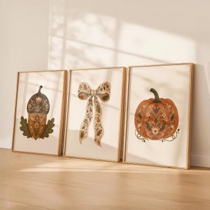 Può includere: Tre stampe artistiche verticali incorniciate in legno chiaro. La stampa a sinistra raffigura una ghianda marrone con motivi floreali e foglie verdi. Quella centrale mostra un fiocco decorativo con motivi botanici. Quella a destra presenta una zucca arancione.