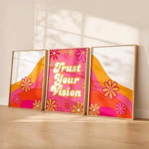Può includere: Set di tre stampe artistiche incorniciate. Ogni stampa presenta un design retrò con uno sfondo ondulato arancione e rosa, accenti floreali e il testo "Trust Your Vision" sulla stampa centrale. Le cornici sono di colore marrone chiaro.