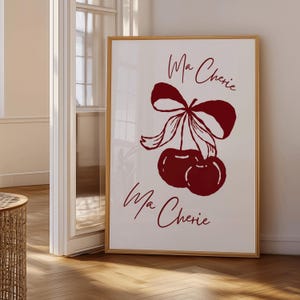 May include: A framed art print featuring a burgundy illustration of two cherries tied with a bow. The words "Ma Cherie" are written above and below the cherries in a cursive font. The frame is a light brown color.
