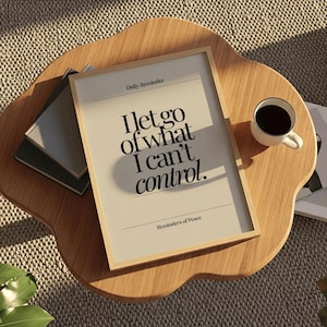 May include: A framed print with the text "I let go of what I can't control" in a minimalist design. The print is on a wooden table with a coffee cup and books.