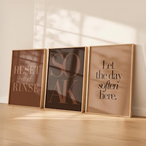 Op de afbeelding: Drie ingelijste kunstafdrukken met goudkleurige frames. De prints bevatten tekst in een schreefletter. De eerste print zegt "RESET and RINSE", de tweede zegt "SOAK" en de derde zegt "Let the day soften here."