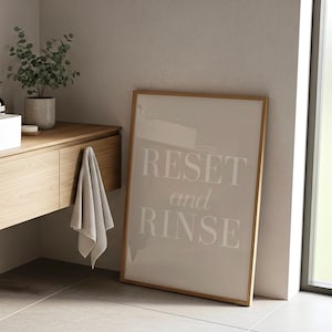 May include: A framed print with the words "RESET and RINSE" in a bathroom setting. The print has a light beige background and a wooden frame. A gray towel hangs from a wooden vanity with a plant in a concrete pot.