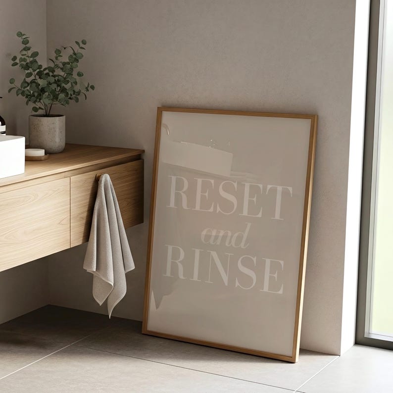 May include: A framed print with the words "RESET and RINSE" in a bathroom setting. The print has a light beige background and a wooden frame. A gray towel hangs from a wooden vanity with a plant in a concrete pot.