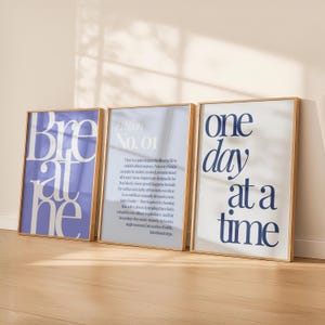 Peut inclure: Trois impressions d'art encadrées avec des citations inspirantes. L'impression de gauche dit "Breathe" en grandes lettres violet clair. L'impression du milieu a du texte et les mots "Edition No. 01". L'impression de droite dit "One day at a time" en bleu marine.