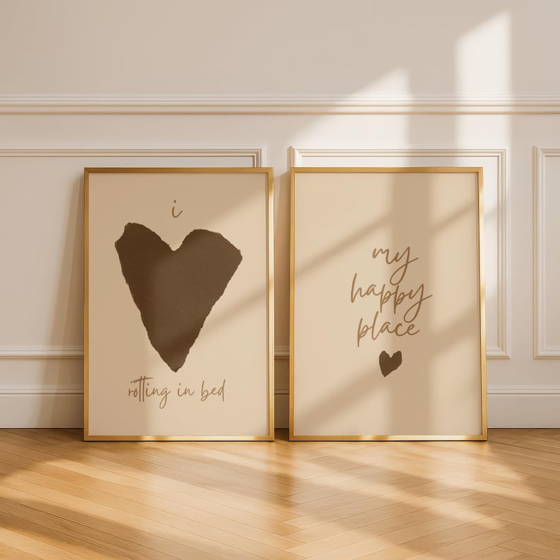 Pode incluir: Duas impress&otilde;es art&iacute;sticas emolduradas com molduras douradas. Uma impress&atilde;o apresenta uma forma de cora&ccedil;&atilde;o marrom escuro com o texto "i rolling in bed". A outra impress&atilde;o tem o texto "my happy place" e um pequeno cora&ccedil;&atilde;o.